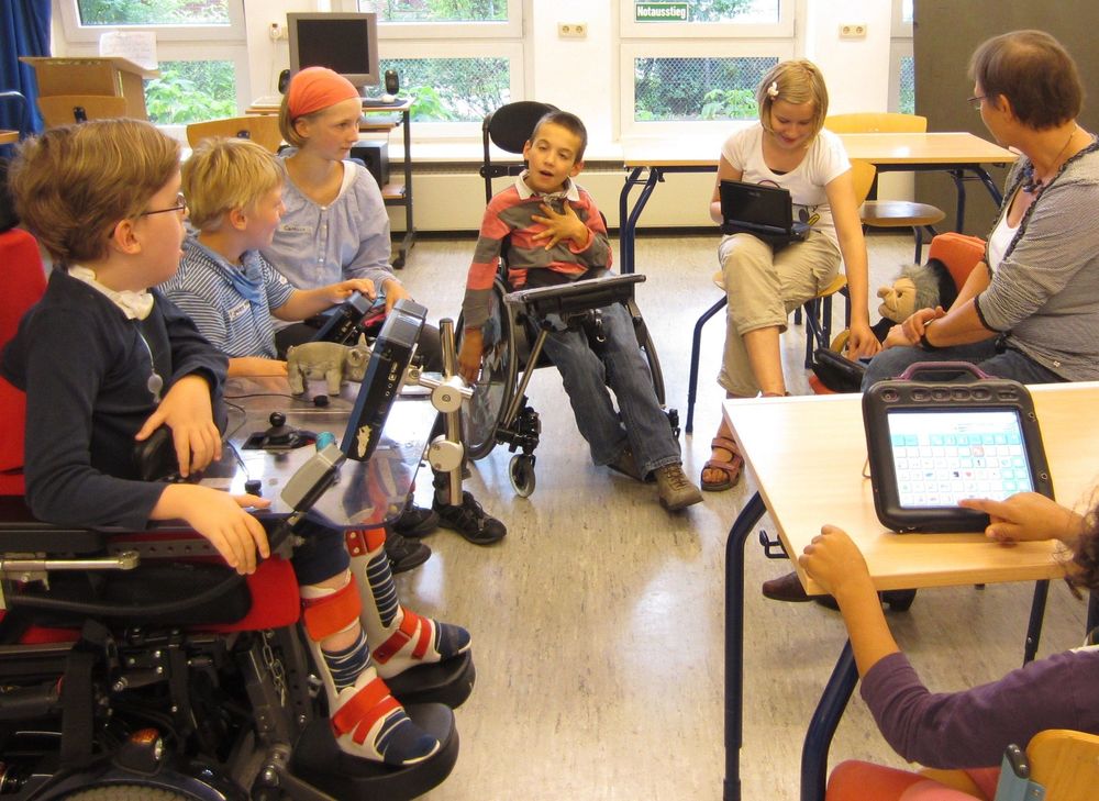 Eine Gruppe von Schüler:innen sitzt im Kreis. Einige im Rollstuhl. Alle kommunizieren mit Hilfe sogenannter Talker (Sprachcomputer).
